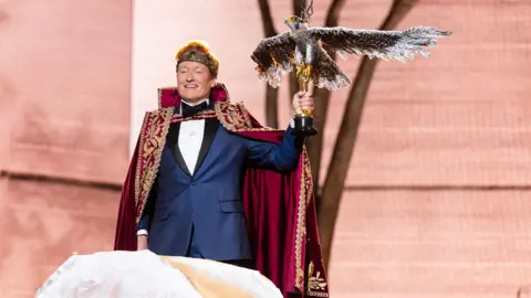Conan O'Brien stands on stage during the Oscars. He's wearing a red and gold cape and gold crown and holding an Oscar statue with a large bird on top.
