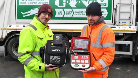 Two refuse collectors standing in front of a bin lorry holding the defibrillators.
They are wearing high-visibility protective clothing and woolen hats.