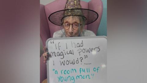 An elderly woman sitting in a pink chair wearing glasses and a witches hat holding up a sign which reads "if I had magical powers I would?" with the answer "a room full of young men".