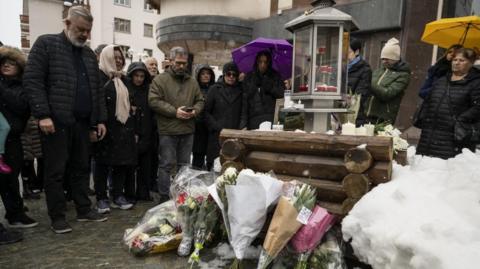 Relatives of victims observe a minute of silence around bouquets of flowers leaning on logs, with a candle-holding container on top, snow around, and with two women with their umbrellas unfurled.