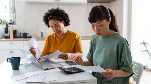 Stock image shows two women in their kitchen looking at bills next to a laptop