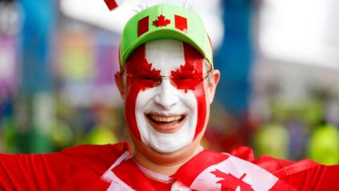 A fan smiles widely in red and white face paint, a red shirt, and a light green hat with a Canadian flag on the front a 