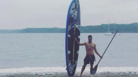 A man stands on a pebble beach beside a large stand-up paddleboard, holding a paddle, with a body of water stretching out behind him. In the background, a sailing boat is anchored near the shoreline, with low hills visible across the water.