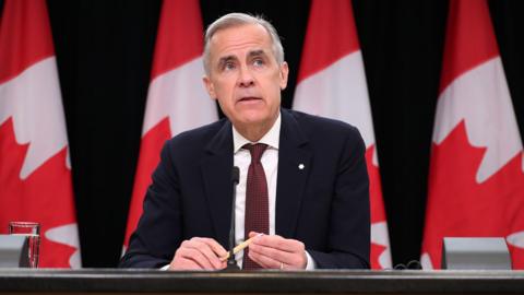 Canada's PM Mark Carney looks to the right of frame while sitting at a press conference briefing table in front of four Canadian flags in Ottowa in May. He wears a black suit and red tie, and holds a pen in his hands with a glass of water beside him on the table.