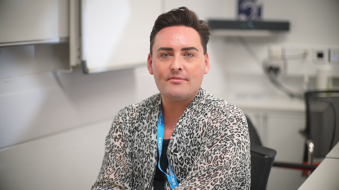 Jamie is sitting in a chair in an office he is wearing a leopard print shirt and black top underneath.
