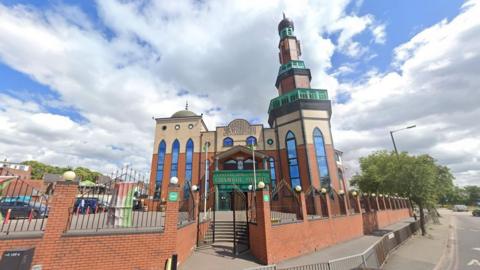 The entrance to a mosque with a green sign that says Central Jamia Mosque Ghamkol Sharif pictured behind a wall.