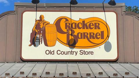 A close up of Cracker Barrel's old logo, with its classic font on a yellow background, on top of one of their stores 
