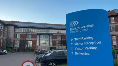 A blue sign at the entrance to Stockton Borough Council's Dunedin House headquarters. It has arrows giving directions to staff parking, visitor parking and reception and for deliveries.