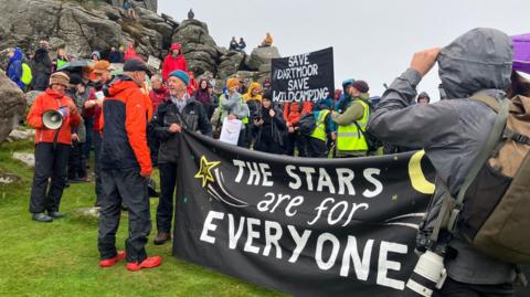 A large group of people wearing camping gear stand in front of Hound Tor. Some are holding a large banner that says 'The stars are for everyone'. In the background another banner says 'Save Dartmoor, save wild camping'.