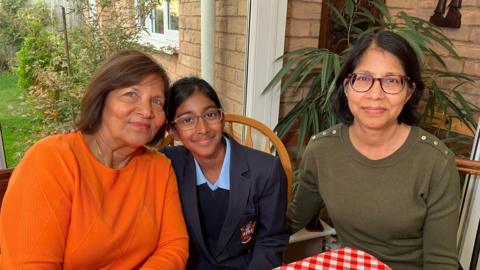 Two women, one in an orange jumper and the other in an olive green coloured jumper sat at a table with a girl dressed in a school uniform sat in the middle.