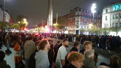 Crowds lining the streets in Dublin City Centre, with the Dublin Spire in the background. It is night time, and the streets are lit.