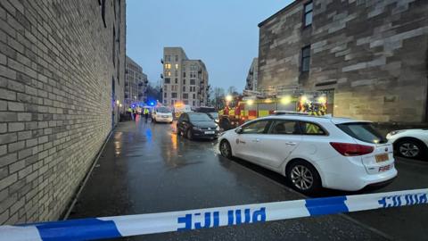 A white car parked behind a line of blue and white police tape. Several other cars are parked on the street outside light-stone coloured buildings. A fire engine is in the middle of the image and in the distance are blue lights and a tower block.