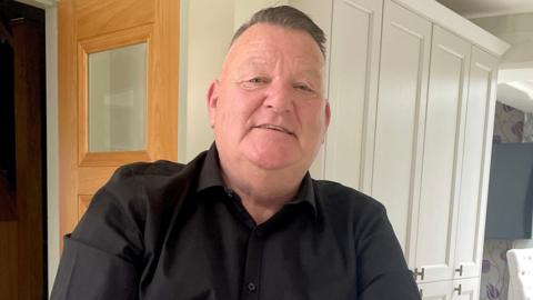 Mark King has short hair and is wearing a black shirt. He is sat in front of a white cupboard and is looking into the camera.