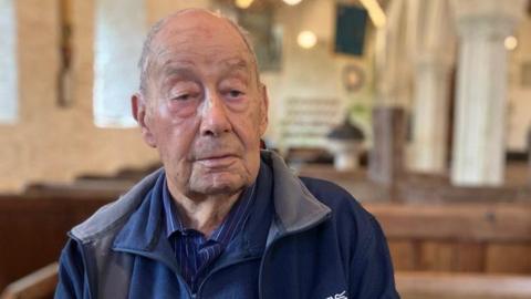 Allan Fisher wears a blue striped shirt, a blue fleece and a blue jacket. He is sitting on a pew in a church