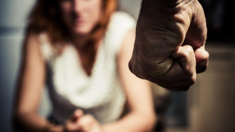 A man's fist coming from the top of the image into shot. In the background is a woman sitting down, who is blurred. 