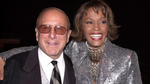 clive Davis and Whitney Houston smiling at the camera. Clive Davis is wearing a black suita nd tie and Whitney Houston is wearing sa jacket covered in silver sequins