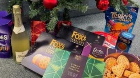 Items on a grey carpet under a Christmas tree, including a box of Heroes chocolates, a bottle of prosecco, Fox's biscuits, a tin of shortbread, Terry's chocolate orange and a tub of Celebration chocolates.