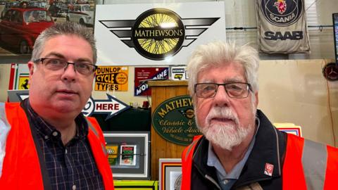 A recent seflie of David with John. David, on the left, wears a blue shirt and a red high vis vest. John, on right, wears a blue shirt, black jacket and a red high vis jacket. Both wear black metal glasses.