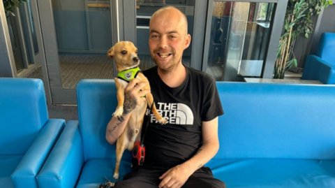 A smiling man wearing a black top and trousers sits on a blue sofa. He is holding a small tan dog which has a yellow collar. 