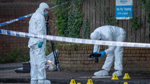 Two people in hazardous material suits stand behind police tape and take pictures