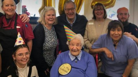 A group of eight people huddled together. In the centre is a woman wearing a pointy, striped birthday hat and a large badge that says it's my birthday. Everybody is smiling towards the camera