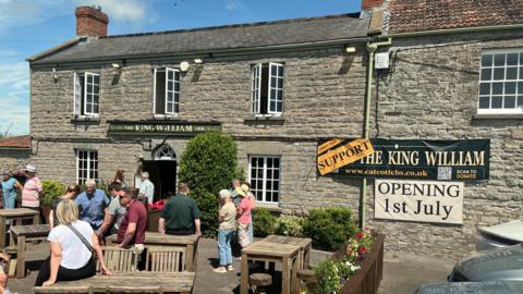 An old grey stone pub is pictured with a sign over the door which says 'The King William Inn'. There are also banners strung across the pub which say 'Support the King William - opening 1st July'. There are several people gathered outside the pub. Most are standing near some wooden tables and chairs.