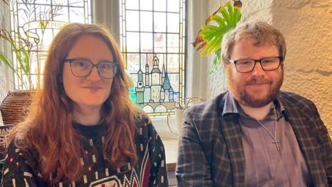 A woman with long ginger hair, black framed glasses and a blue patterned jumper next to a man with ginger hair, a ginger beard and black framed glasses in a blue shirt and blue chequed jacket in front of a stained glass window