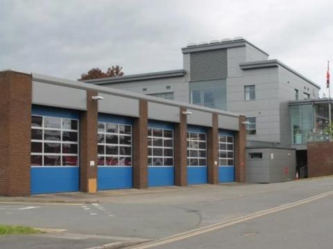 A fire station pictured from the road outside. A high appliance bay can be seen, with four large doors that are blue in between brick pillars. A large grey building is on the right.