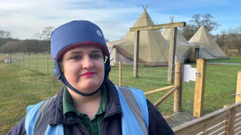 Anna wearing a blue hat. There is a tee-pee on farm land behind her. She is wearing a blue hi-vis jacket and a warm fleece.