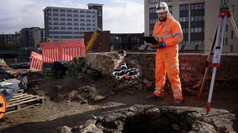 A man in an orange boiler suit holding a screen as he stands next to a well on a building site in Sheffield. He is operating a drone.