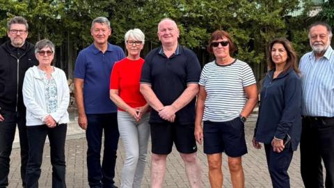 Residents Paul Briggs, with short grey hair and glasses and dressed all in black; Sheena Briggs, with short grey hair, sun glasses, cream hoodie and black trousers; Fred Hood, with short grey hair, blue t-shirt and black trousers; Debbie Hicks, with short white hair, glasses, black and white striped trousers and red top; Malcolm Layford, with white hair, wearing black shorts and t-shirt; Jean Sutcliffe, with short brown hair and sunglasses, wearing black shorts and a black and white striped t-shirt; Jas Chera, with long brown hair, wearing a navy blue shirt and black trousers and Jinder Chera, with white hair and beard, wearing black trousers and blue and white striped shirt.