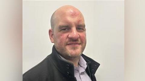 Head and shoulders of David Heap. He has a bald head and stubble and is wearing a dark coat with a stand up collar