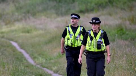 A male and female police officer walking on a track through a grassy area. They are wearing hi-viz vests over their black uniforms.