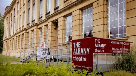 The exterior of a theatre, it is sand coloured stone building and there is a metal ramp leading to the entrance as well as a burgundy sign that reads "The Albany Theatre".