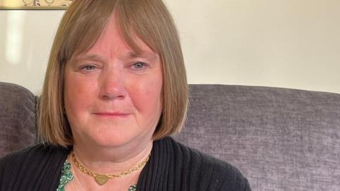 A woman sits on a sofa looking towards the camera and frowning. She has light brown hair, in a bob, and wears a black cardigan, a green top and a gold necklace.