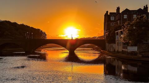 Sunset over a bridge over a river, the sun is reflected on the water.