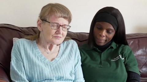 Woman in her 60s sits left of frame wearing a blue v-neck jumper, her carer is sitting on the right