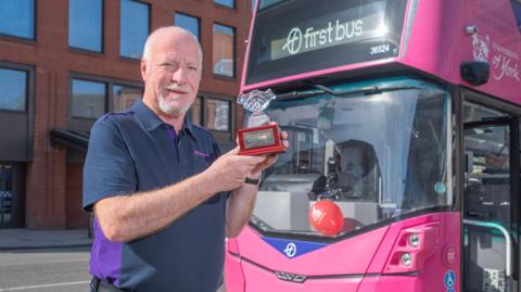 A man standing outdoors on a street in front of a bright pink bus. The bus has a digital display on the front that reads “first bus”, along with a fleet number 36524. The bus also carries branding for the University of York near the upper right corner of the windshield. A red balloon is visible inside the bus near the front window.