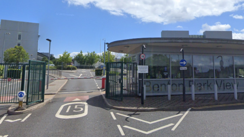 The entrance to the Benton Park View building. There is a green fence, red and orange barrier on the road and a glass fronted building with a security guard sitting inside. A grey building can be seen in the background.