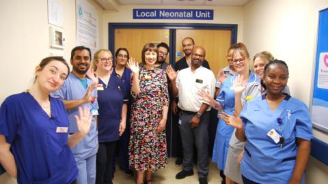 Lots of hospital staff standing in a hospital corridor. They have their hands up waving and smiling at the camera. 