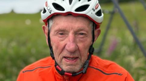 Eddie looks directly at the camera with a determined look. He's wearing a white cycling helmet and orange top.