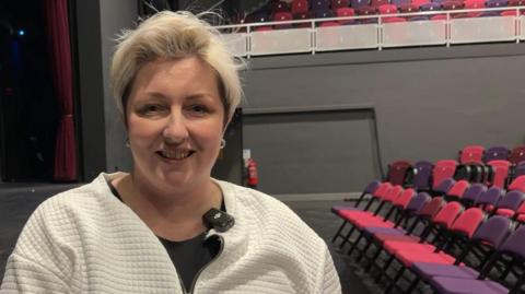 A woman with short blond hair stands in a large theatre auditorium with rows of red and purple seats to the right and a stage curtain to the left. She is wearing a white cardigan over a black top.