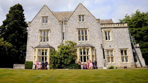 An external photo of Larkbeare House with wedding guests standing in front of it, a lawn in the foreground and a large fir tree to the left of the house