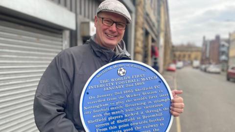 A smiling man with a flat cap and glasses holds a blue plaque in front of a series of railway arches