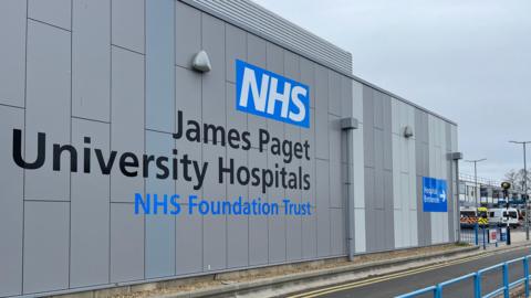 The outside of the James Paget University Hospital building. The grey outside of the building has black and blue writing on it which says NHS James Paget University Hospitals NHS Foundation Trust.