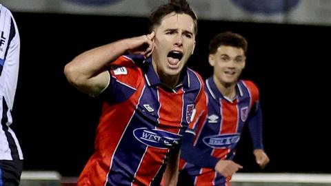 Joe Haigh in red and blue striped Dagenham shirt runs away celebrating a goal by pointing to his head as a team-mate tries to catch up with him