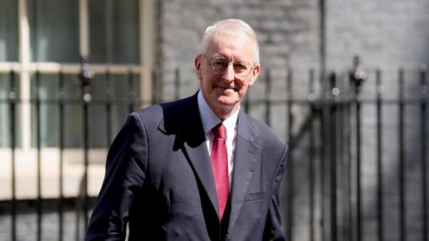 Hilary Benn outside 10 Downing Street. He has grey hair and glasses and is wearing a dark suit with a red tie.