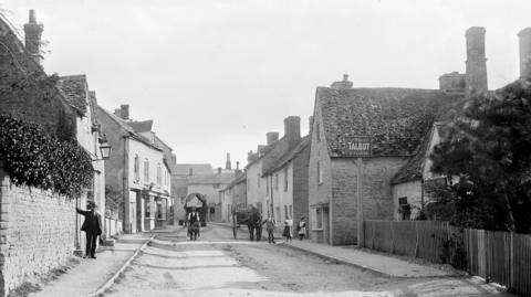 An historical photograph of a wide street in the town of Charlbury, Oxfordshire, taken around the end of the 18th Century. A man can be seen leading a horse and cart, and another leaning against a wall.  