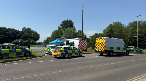 Four police cars and two police vans parked at the entrance of a park on a sunny day.