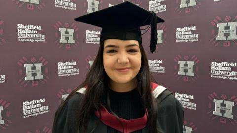 Ms McGoldrick stands before a Sheffield Hallam University banner, wearing her graduation gown. She has long dark brown hair and smiles at the camera.